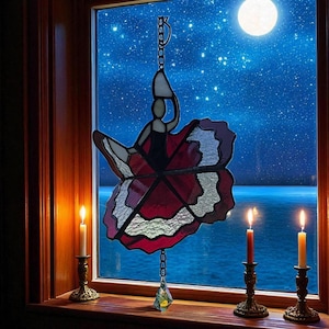 May include: A stained glass window decoration depicting a dancer with a red and white skirt, hanging in front of a night sky with a full moon. Two lit candles in brass holders sit on the windowsill, adding a warm glow.