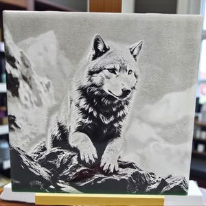 May include: Black and white artwork of a wolf sitting on a rocky surface, with a mountainous background. The wolf is detailed with fur and facial features. The artwork is displayed on a wooden easel.
