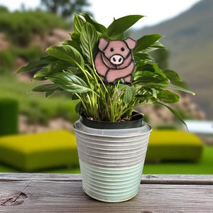May include: A potted plant with a decorative pig-shaped plant stake. The pig is pink with black outlines. The pot is a light blue and white striped design. The background is a blurred outdoor scene.