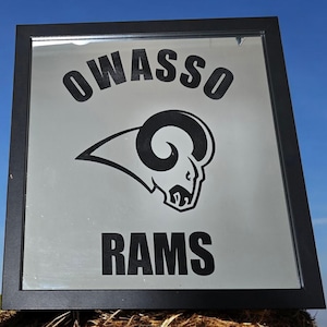 May include: A black framed mirror with a silver background. The mirror has a black and white graphic of a ram's head with the text "OWASSO RAMS" above it.