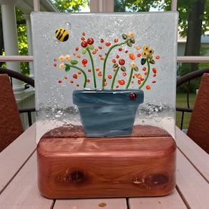 May include: A decorative glass panel featuring a blue flower pot with colorful glass flowers and a ladybug. A yellow and black bee is on the left side of the panel. The panel is mounted on a wooden base.