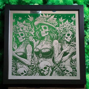 May include: A framed print depicting three skeletons wearing floral headbands and holding marijuana leaves. The print is set against a green background and has a white border.