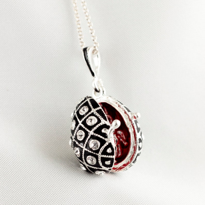 May include: A silver necklace with a pendant shaped like an egg. The egg pendant is black with a diamond pattern and clear crystal accents. The inside of the egg is red. The pendant is open, revealing the red interior.