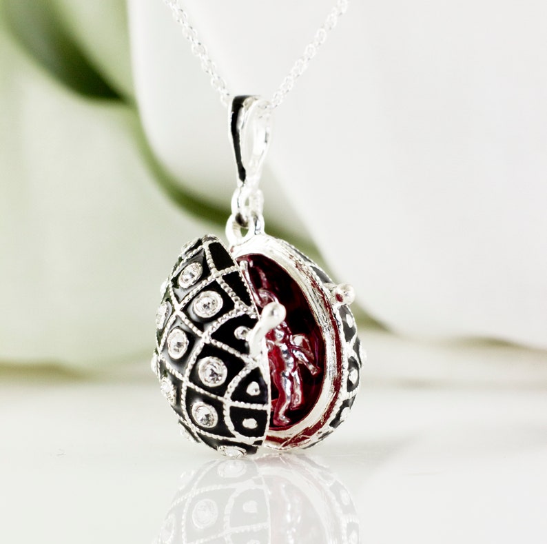 May include: A silver necklace with a pendant shaped like an egg. The egg is black with a geometric pattern and clear crystals. Inside the egg is a red interior with a small figure. The pendant is open, revealing the interior.