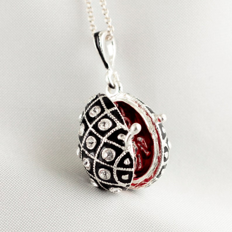 May include: Silver-tone necklace with a black and white patterned egg-shaped pendant. The pendant opens to reveal a red interior and is embellished with clear crystals. The necklace is displayed on a white background.