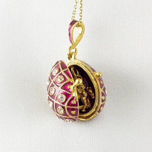 May include: A gold-colored egg pendant necklace with a hinged opening. The egg is decorated with a red enamel pattern, clear crystals, and a small gold-colored figurine inside. The necklace hangs on a delicate gold chain.