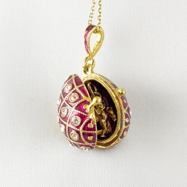 May include: A gold-colored egg pendant necklace with a hinged opening. The egg is decorated with a red enamel pattern, clear crystals, and a small gold-colored figurine inside. The necklace hangs on a delicate gold chain.