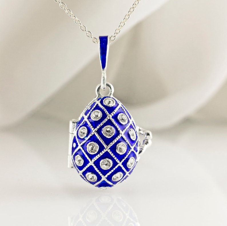 May include: A silver necklace featuring a blue enameled egg pendant. The egg-shaped pendant has a diamond pattern with clear crystal accents. The pendant opens to reveal a small compartment. The necklace is a delicate piece of jewelry.