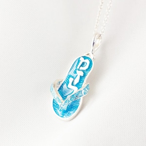 May include: A silver necklace with a blue and white flip-flop pendant. The pendant has a detailed design with the word "DTS" in white. The flip-flop is a vibrant blue color and is suspended from a delicate silver chain.