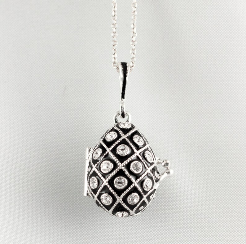 May include: Silver-tone teardrop pendant necklace. The pendant features a black enamel design with a diamond pattern and clear crystal accents. The pendant opens to reveal a small compartment. The necklace hangs on a delicate silver chain.