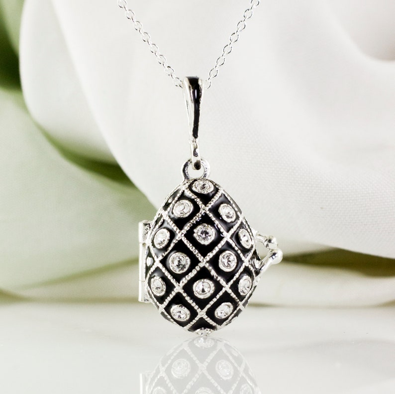 May include: A silver-toned egg-shaped pendant necklace with a black enamel grid pattern and clear crystal accents. The pendant opens to reveal a small compartment. The necklace hangs on a delicate silver chain.