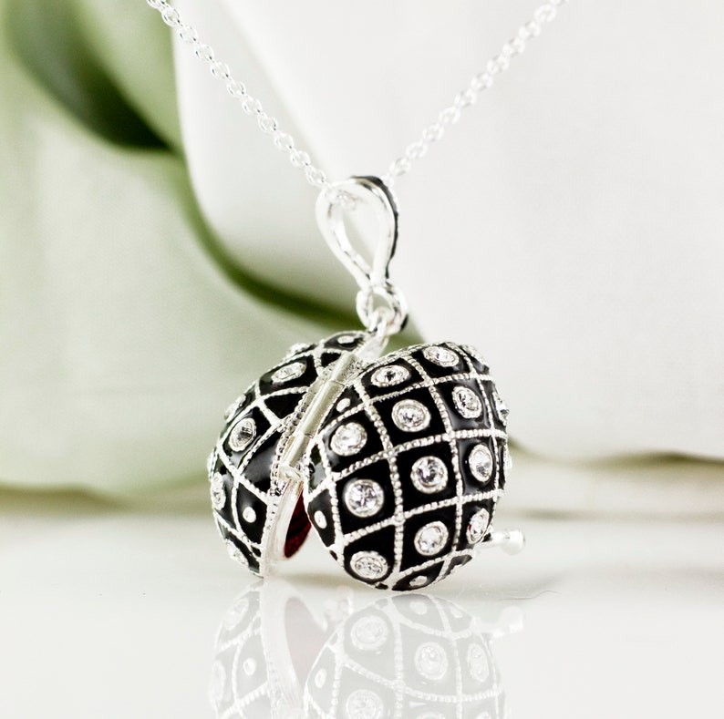 May include: A silver necklace with a pendant shaped like an egg. The egg pendant is black with a grid pattern and clear crystal accents. The pendant opens to reveal a red interior.