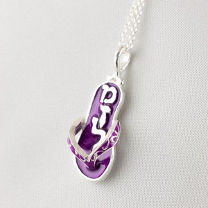 May include: A silver chain necklace with a purple flip-flop pendant featuring the Hebrew word "Shalom" in white lettering.