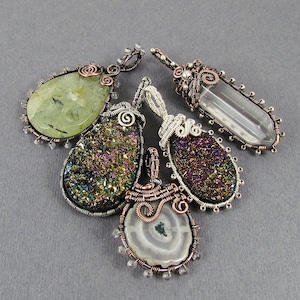 May include: Five wire-wrapped pendants featuring a variety of gemstone cabochons. The pendants showcase a range of colours and textures, including green, purple, and white. The wire wrapping is crafted in copper and silver.
