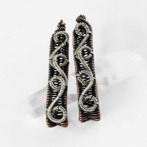 May include: Two handcrafted wire-wrapped earrings. Each earring is rectangular with a woven black and copper base. A silver-colored wire design decorates the front. The earrings are displayed on a white surface.