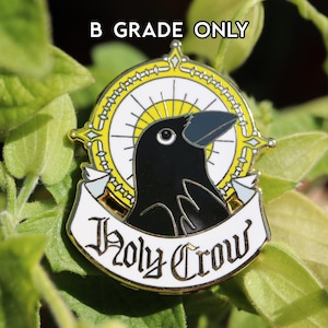 Könnte beinhalten: Eine schwarze und gelbe Emaille-Anstecknadel mit einer Krähe mit Heiligenschein und dem Text "Holy Crow".