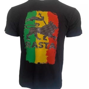 Puede incluir: Camiseta negra con un gráfico de león Rasta verde, amarillo y rojo con diamantes de imitación. El gráfico incluye la palabra "RASTA" en diamantes de imitación.