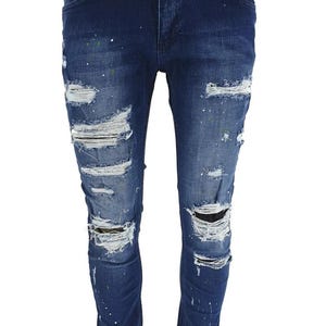 Puede incluir: Vaqueros azules de denim con detalles desgastados y salpicaduras de pintura blanca. Los vaqueros tienen múltiples roturas y desgarros en las piernas.
