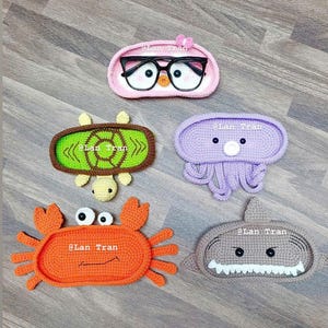 May include: Five crocheted animal-shaped eyeglass holders. The holders are shaped like an owl, a turtle, a jellyfish, a crab, and a shark. Each holder has a small, oval-shaped space for holding eyeglasses.