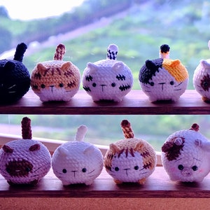May include: A collection of 12 crocheted cat and pig plushies in various colors and patterns. The plushies are arranged on a wooden shelf with a window in the background.