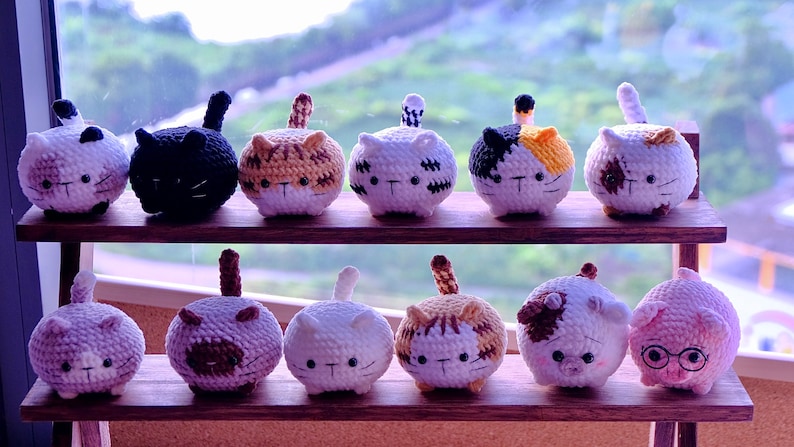 May include: A collection of 12 crocheted cat and pig plushies in various colors and patterns. The plushies are arranged on a wooden shelf with a window in the background.