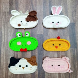 Op de afbeelding: Zes gehaakte dierenmanden. De manden zijn in de vorm van een koe, konijn, kikker, kuiken, kat en varken. De manden zijn allemaal wit met verschillende gekleurde accenten.