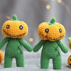 May include: Two handmade crochet pumpkin-headed figures with green bodies and stitched details. The pumpkins are orange with black stitched smiles and cross-shaped eyes. Additional pumpkins are in the background, suggesting a Halloween theme.