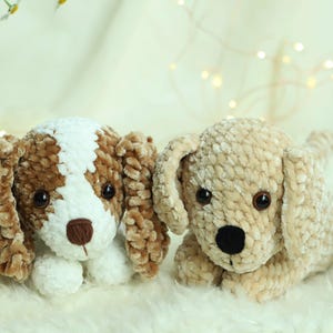 Könnte beinhalten: Zwei gehäkelte Plüsch-Hunde-Spielzeuge. Eines ist braun und weiß mit einem weißen Gesicht und braunen Ohren. Das andere ist beige mit einem beigen Gesicht und Ohren.