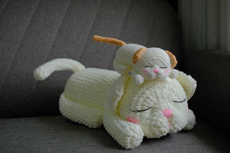 May include: A handmade, off-white crochet dog and puppy plush toy. The larger dog has closed eyes, pink accents on the nose and cheeks, and a curled tail. The smaller puppy rests on top, also with closed eyes and pink accents.