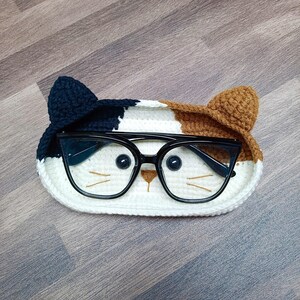 May include: A crocheted cat-shaped glasses holder with black, white, and brown yarn. The holder has black eye details and a pair of glasses resting inside.