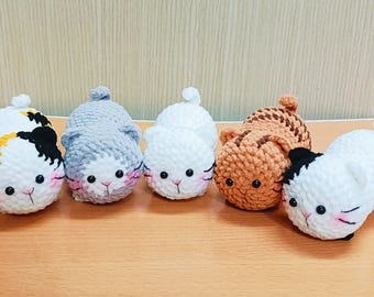 May include: Five crocheted cat plushies in different colors and patterns. The plushies are all round and have black eyes and pink cheeks. They are arranged in a row on a wooden surface.
