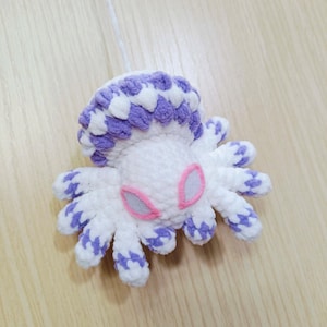 May include: A white crocheted octopus with purple and white stripes and pink eyes. The octopus is hanging from a white string.