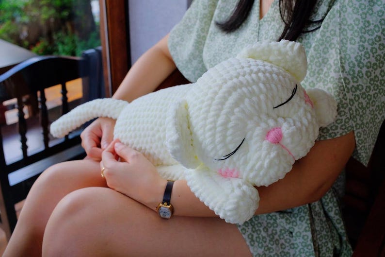 May include: A large, white, crocheted lamb plush toy with closed eyes and pink accents on the cheeks. The lamb has a long tail and floppy ears. The toy is soft and cuddly, perfect for a child's bedroom or nursery.