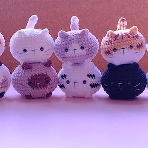 May include: Six crocheted cat plushies stacked in pairs. The plushies are white, brown, black, and orange with black eyes and whiskers.