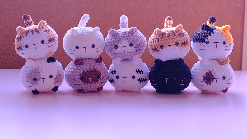 May include: Six crocheted cat plushies stacked in pairs. The plushies are white, brown, black, and orange with black eyes and whiskers.