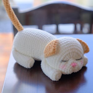 May include: A handmade, crocheted cat toy in a sleeping pose. The cat is primarily white with light brown ears and a tail. It has closed eyes, pink cheeks, and a pink nose. The toy is lying on a dark blue surface.