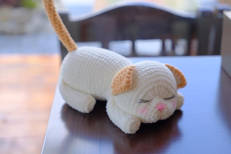 May include: A handmade, crocheted cat toy in a sleeping pose. The cat is primarily white with light brown ears and a tail. It has closed eyes, pink cheeks, and a pink nose. The toy is lying on a dark blue surface.