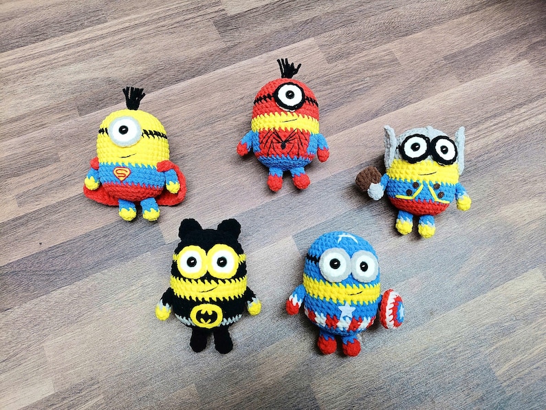 May include: Five handmade crochet Minion-style figures, each with a superhero theme. The figures are yellow, blue, red, and black, with details like a Superman logo, Spider-Man web, Thor's hammer, and Captain America's shield. They are arranged on a wooden surface.