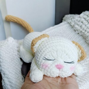 May include: A handmade white crochet cat toy with tan accents on the ears and tail. The cat has closed eyes, pink cheeks, and a pink nose. The toy is lying on a white surface.