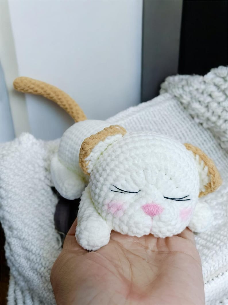 May include: A handmade white crochet cat toy with tan accents on the ears and tail. The cat has closed eyes, pink cheeks, and a pink nose. The toy is lying on a white surface.