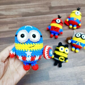 May include: A collection of handmade crochet Minion figures. One Minion is blue, yellow, and white, holding a shield. Other Minions are in various colors and designs, including red, yellow, and black. The figures are on a wooden surface.