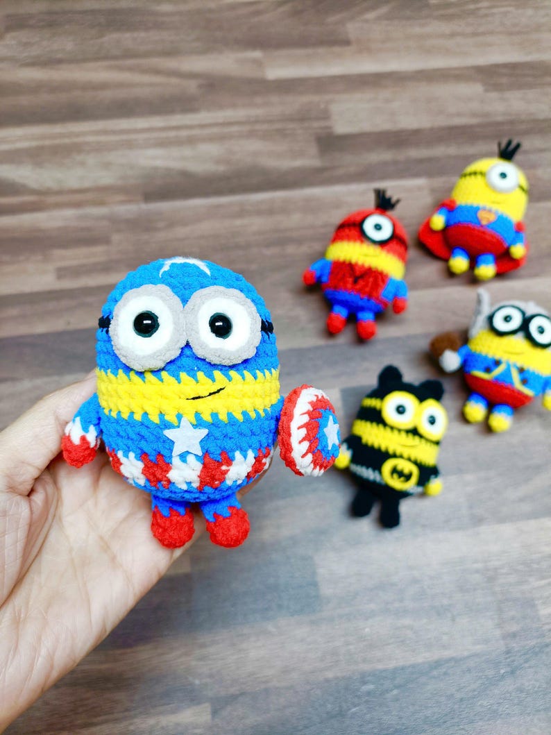 May include: A collection of handmade crochet Minion figures. One Minion is blue, yellow, and white, holding a shield. Other Minions are in various colors and designs, including red, yellow, and black. The figures are on a wooden surface.