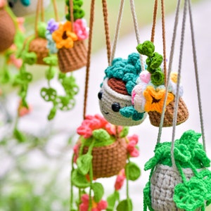 May include: A collection of crocheted hanging planters with various colors and textures. The planters feature different types of flowers and greenery, including succulents, leaves, and flowers. The planters are suspended from a string with clothespins.