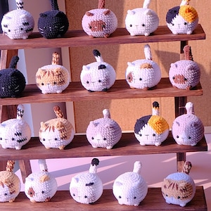 Può includere: Un espositore in legno con 20 peluche di gatto all'uncinetto in vari colori e motivi. I peluche sono disposti su tre file sullo scaffale.