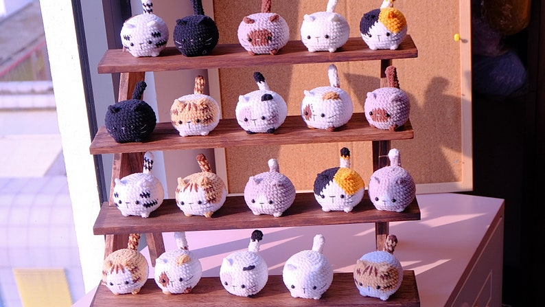 May include: A wooden shelf display with 20 crocheted cat plushies in various colors and patterns. The plushies are arranged in three rows on the shelf.