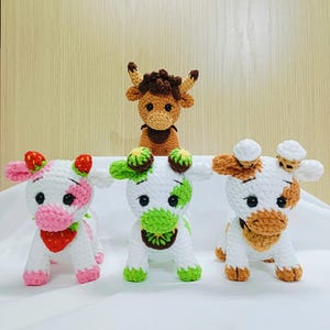 May include: Four crocheted stuffed cows, two with white and brown spots, one with white and pink spots, and one brown cow. The cows with spots have different coloured accents on their necks, one with green, one with pink, and one with brown.