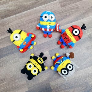May include: Five handmade crochet Minion figures, each dressed as a superhero. The figures are yellow, blue, red, and black, with details like a Captain America shield, Spider-Man web design, and a Batman symbol. They are arranged on a wooden surface.