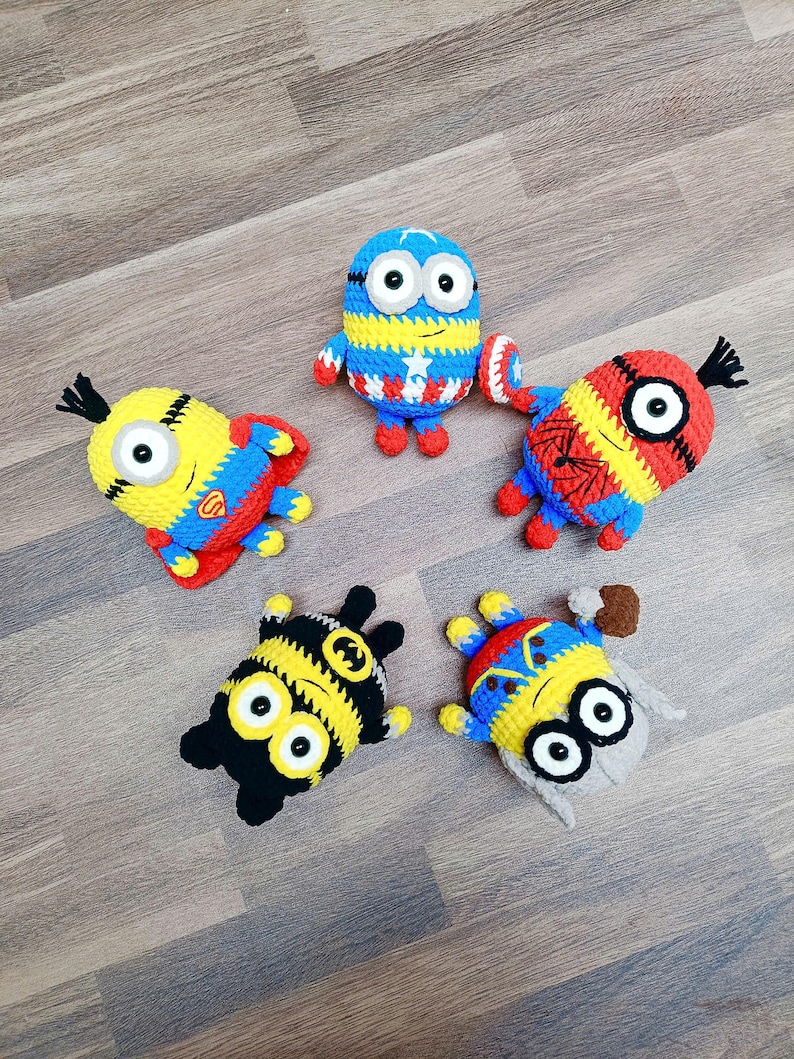 May include: Five handmade crochet Minion figures, each dressed as a superhero. The figures are yellow, blue, red, and black, with details like a Captain America shield, Spider-Man web design, and a Batman symbol. They are arranged on a wooden surface.