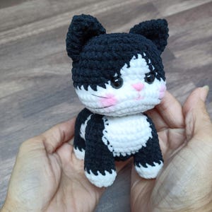 Könnte beinhalten: Eine handgefertigte Häkelkatze in Schwarz und Weiß. Die Katze hat schwarze Ohren, einen schwarzen Kopf und ein weißes Gesicht mit rosa Wangen. Der Körper ist schwarz und weiß, und die Katze wird in zwei Händen gehalten.