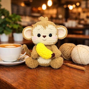 Pode incluir: Uma boneca de macaco de crochê em tons de marrom e creme, segurando uma banana amarela. O macaco tem olhos pretos e bochechas rosadas. Uma xícara de café e novelos de lã ao fundo.